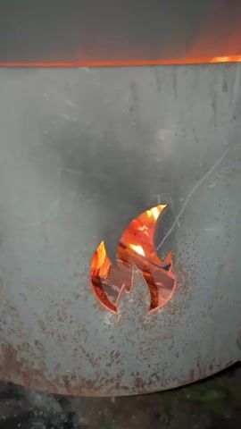 This video shows a close-up of a metal stove with a flame symbol cut out of its door. Inside the stove, flames are visible, reflecting through the cutout. The stove is outdoors, with grass and debris on the ground around it. The flames appear to be strong and are licking up the sides of the stove. The metal of the stove is aged and shows signs of rust and wear. The lighting suggests it is either dusk or nighttime.