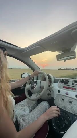 A blonde woman drives a white Fiat 500 convertible on a road with a field of wheat on the right. The sun is setting, casting a warm glow on the sky and reflecting on the car's dashboard. The woman is wearing a white dress and has her hands on the steering wheel and gear shift. The sunroof is open, and the interior of the car is visible, including the dashboard, steering wheel, and gear shift. The video is shot from inside the car, looking towards the driver and the road ahead. The overall mood is serene and romantic.