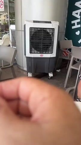 A close-up, first-person perspective video shows a hand holding a single purple flower between the thumb and index finger. The flower is in focus in the foreground. In the blurred background, a white and gray evaporative cooler is visible, along with a white pillar, and part of a green sign with white lettering that appears to say "HAND FO...". The setting appears to be outdoors or in a covered outdoor area, possibly a cafe or restaurant. The hand is positioned to the right of the frame, and the flower is held towards the center. There are no other visible people or distinct actions occurring. The lighting suggests it is daytime.