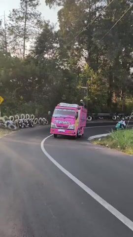 The video opens with a bright pink, custom-decorated pickup truck driving on a paved road. The truck has elaborate graphics and lettering in various colors, including yellow, blue, and white, along its sides and the back. A person is standing in the bed of the truck, appearing to be filming or directing. The road is curved, with a white line marking the lane. On the left side of the road, there are a series of tires stacked and arranged as a barrier, and beyond that, trees and foliage. On the right side of the road, a motorcycle rider on a scooter is visible, following behind the truck. The environment appears to be rural or suburban, with power lines and poles visible. The scene shifts to a closer view of the truck's graphics, showcasing more of the detailed artwork and lettering. The camera then pans to the rear of the truck, revealing more of the decoration and the rider in the bed. The perspective moves to follow the truck and scooter from behind as they continue on the road, which winds through a landscape with trees, grass, and some buildings in the distance. The sky is visible, with some clouds. The video ends with a shot of the truck and scooter turning a corner on the road.
