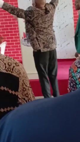The video depicts a man in a brown-patterned blazer and dark pants standing on a red-carpeted platform addressing an audience. He holds a microphone and gestures with his right hand. Behind him is a white wall with a red brick pattern at the base and a logo. The audience is visible from the back, with one person wearing a black and white patterned shirt and another in a blue shirt. The man on the platform is bald and appears to be speaking or singing.