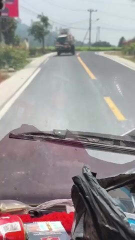 The video shows a view from inside a vehicle, looking out through the windshield. The windshield is dirty and has windshield wipers. In the distance, a truck loaded with what appears to be scaffolding or building materials is driving on a paved road with a yellow dividing line. The truck is moving away from the camera and turning to the right. To the right of the truck, a motorcycle with two people on it is also traveling on the road. The environment appears to be rural, with greenery and trees visible on the sides of the road. There are also power lines and utility poles in the background. The sky is bright, suggesting it is daytime. The foreground shows some items inside the vehicle, including what looks like packaging and a red object.