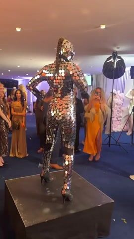 A person wearing a full-body, mirrored-disc suit stands on a black platform surrounded by onlookers. The suit covers the person from head to toe, including a hood, with reflective silver discs. The person wears matching high-heeled boots. The platform is in the center of a large room with a blue carpet, surrounded by people in formal wear.  Some of the onlookers are taking pictures or videos with their phones, while others are conversing. A photographer with a professional camera is positioned to the right of the platform. Overhead lighting is visible, as well as some decorations. The atmosphere appears to be celebratory. Confetti is scattered on the floor.