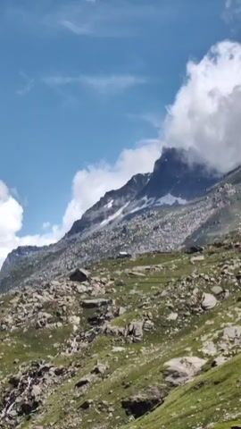 The video pans across a mountainous landscape under a bright blue sky with scattered clouds. The foreground consists of a rocky, grassy slope with a small, dark-colored structure situated partway up. Further back, rugged, rocky mountain slopes rise sharply, with patches of snow visible near the peaks. The camera movement reveals a deep valley with winding roads or paths and more majestic, snow-capped mountains in the distance. The overall impression is of a vast, natural, and somewhat remote alpine environment.