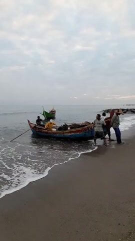 The video shows a group of men bringing a small wooden boat ashore on a sandy beach. There are five men visible. One man is in the boat using an oar, and four others are standing in the shallow water, pushing and pulling the boat onto the beach. The boat is filled with fishing nets. In the background, another small, green boat is anchored in the water. The sky is overcast, and the sea is calm with small waves lapping at the shore. The beach is mostly sandy with some seaweed or debris scattered along the shoreline. In the far background, there are what appear to be rocks or breakwaters.