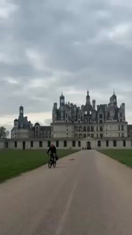 Here is a description of the video:

The video shows two individuals riding bicycles on a gravel road leading toward a grand chateau under an overcast sky. The chateau is large and elaborately designed, with multiple spires and decorative elements. The road is bordered by a grassy area on either side, and the camera follows the cyclists as they approach the chateau. The cyclists are wearing casual clothing, and one of them has a backpack.