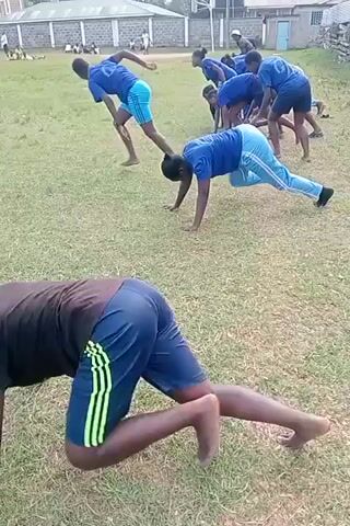 The video shows a group of people, possibly a sports team, practicing on a grass field. Most of them are wearing blue shirts and blue or light-colored pants or shorts. They are engaged in exercises that involve running and doing push-ups. A rugby ball is lying on the grass near the center of the field. In the background, there is a multi-story building with a brick facade, other people sitting or standing, and a fence. The camera appears to be held by someone who is also participating in the exercises, as it moves with them.