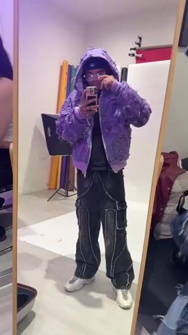 A full shot of a man taking a selfie in front of a mirror, with a woman sitting out of focus in the background on the left. He is wearing a purple, heavily textured hooded jacket, a black undershirt, black and grey baggy cargo pants with chains, and white sneakers. He's also wearing glasses, a hat, and a chain necklace. He's holding a smartphone up to take the picture. The mirror has a thin wooden frame. The background shows a studio space with a white backdrop, lighting equipment, and color rolls propped against the wall. There are some items, including a red jacket, in the corner on the right side of the mirror. The room appears to be well-lit, though there seems to be purple lighting.