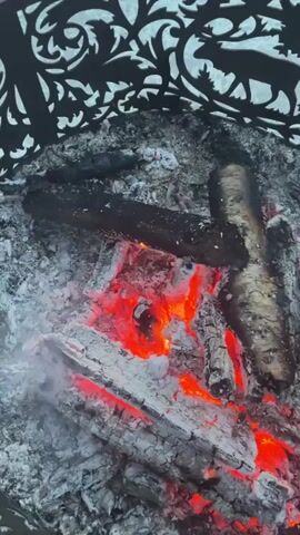 The video shows a backyard barbecue setup. A metal fire pit with decorative cutouts contains glowing embers and charred wood. The camera pans up and reveals a patio area with a yellow folding chair.  A block wall is visible in the background, and a purple covering is placed over some of the blocks. A barbecue grill is next to the wall with sausages cooking on it. There is a table with a white tablecloth and a bottle of liquor on it. The yard is grassy, and trees surround the area.