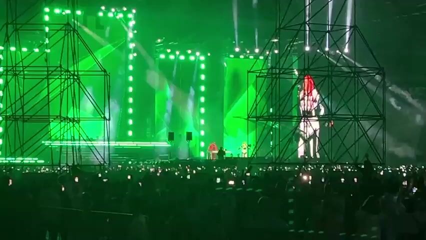A concert is taking place on a large stage. The stage is lit with bright green and white lights, creating beams of light that shine into the audience. In the background, a large screen displays animated visuals, predominantly in green. There are scaffolding structures on either side of the stage. On the stage, several figures can be seen, with one person in a red outfit standing in the center, seemingly performing. The audience is large and is visible in the foreground, with many people holding up their phones, likely recording or taking pictures. The overall atmosphere appears to be energetic and vibrant.