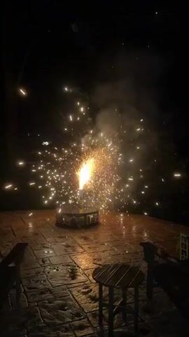 The video shows a firework exploding on a patio at night. Sparks are flying in all directions from the firework, which is sitting in a metal base. The patio is made of stone and appears to be wet. There are trees in the background, and the sky is dark. Two chairs and a small table are visible on the patio.