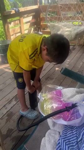 A young boy wearing a yellow t-shirt and blue Crocs is standing on a wooden deck, using a black hand pump to inflate a clear plastic toy that is filled with pink and yellow confetti. The boy is bending over the toy, focusing on the inflation process. In the background, there is a wooden railing and some greenery. A white furry blanket and a blue and white striped towel are visible in the foreground. The camera is positioned at eye level with the boy.