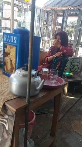 A woman wearing a headscarf and a red patterned shirt is sitting on a chair and looking to the side. In front of her, on a wooden table, is a silver teapot, two glasses of water on a red tray, and a blue container with the word "RAOS" and food items inside. A metal pole is in the foreground. The environment appears to be outdoors, possibly a street food stall or market, with a corrugated metal roof overhead and structures in the background.