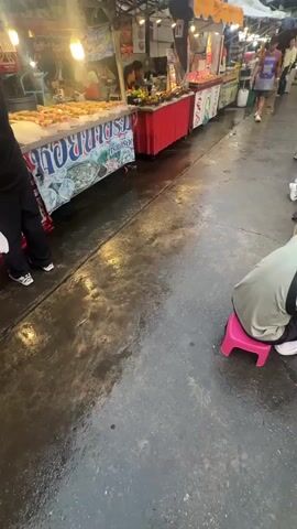 The video shows a street market scene. In the foreground, a person is kneeling on a pink stool, facing away from the camera, and appears to be looking at items spread out on the ground. The ground is wet and reflects the lights from the market stalls. To the left, a market stall displays food items on ice, with a sign written in a foreign language. Behind this stall, other market stalls are visible, with vendors and customers. People are walking along the street, and some are browsing at the stalls. The lighting suggests it is either dusk or a dimly lit area. The overall atmosphere is that of a busy outdoor market.