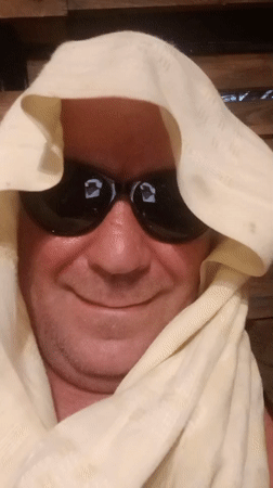 Smiling selfie in sunglasses and headscarf, blurred outdoor backdrop.
