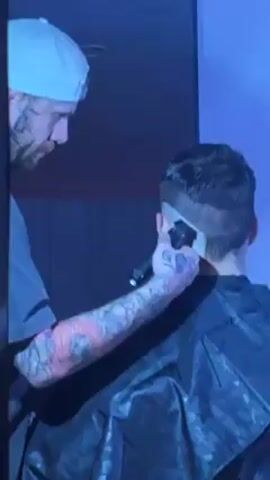 A man with tattoos and a backwards baseball cap is giving a haircut to a person wearing a black barber’s cape. The man is wearing a gray t-shirt with the words “THIS IS SOJI” on the back.  He is using an electric razor to cut the hair on the back of the person’s head.  The person getting their haircut is obscured by the barber’s cape and the camera angle.  After he finishes with the electric razor, the man walks out of frame. He holds a pink comb behind his back. The environment is dimly lit, with the colors in the scene appearing to be blue.