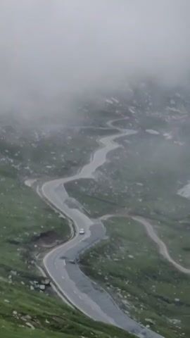 A white car navigates a winding mountain road that cuts through a grassy, rocky landscape. The road is paved and has white lines marking the lanes. The surrounding terrain is steep and covered in green grass and scattered rocks. A thick fog or mist hangs in the upper portion of the frame, obscuring the distant landscape and creating a sense of altitude and atmospheric conditions. The car is traveling uphill, appearing to be making a turn. The overall scene conveys a sense of isolation and the journey through a challenging natural environment.