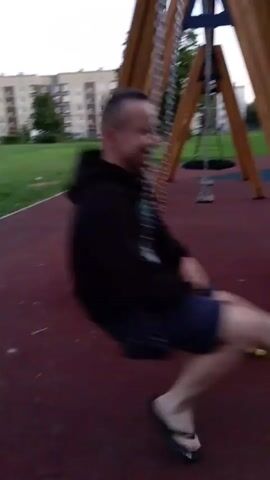 A person is on a swing set in a park. They are wearing a black hoodie and blue shorts, with flip-flops on their feet. The person is swinging back and forth. There are juice boxes and snacks on the ground near the swing set. In the background, there are apartment buildings and trees. The video is shaky, suggesting it was filmed by someone holding the camera.
