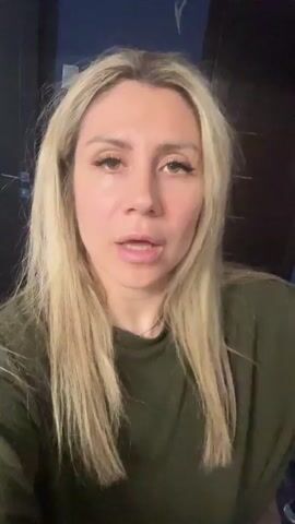 The video shows a blonde woman with light makeup and long straight blonde hair. She is wearing a dark green ribbed t-shirt. In the first few seconds, she is seen pulling on her hair and her mouth is open as if speaking. Her eyes are looking directly at the camera. The background is dark, with hints of blue on the wall and a dark brown door or cabinet to her right. The lighting is somewhat dim, casting shadows on her face.