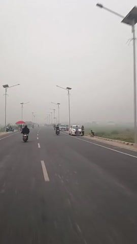 The video shows a straight, paved road with a white solid line down the center. The sky is overcast and hazy. There are solar-powered streetlights on both sides of the road. People are riding motorcycles and standing on the side of the road, some near parked cars. A red umbrella is visible on the left side of the road. In the distance, more vehicles and people are present. The overall environment appears to be a rural or suburban area with limited visibility due to the haze.
