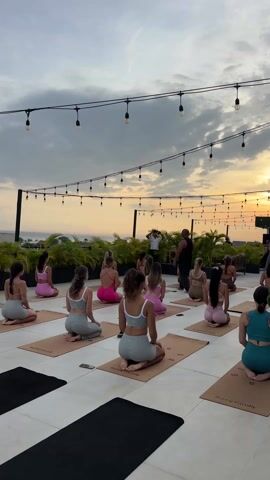 The video shows a yoga class taking place on a rooftop at sunset. There are about 15 women sitting on yoga mats, facing a male instructor who is demonstrating a breathing exercise. The women are all wearing sports bras and leggings or shorts, in a variety of colors including gray, pink, and teal. The instructor is wearing a black tank top.  A second man stands in the background, partially obscured. Strings of light bulbs are strung above the group. In the background, there is a view of the ocean and greenery. The mats are tan with black writing on them that says “Sunny Family”. There are a few black mats on the left side of the frame.