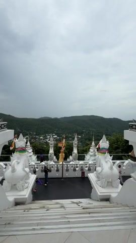 The video shows a view from a set of white marble stairs looking out over a landscape. In the foreground, on the left and right sides of the stairs, are white decorative statues of mythical creatures, possibly lions or dogs, with intricate details. There are also white, ornate structures that appear to be part of a temple or shrine. In the middle ground, there is a balcony-like area with more white decorative elements and several small golden flags on poles. A few people, dressed casually, are standing on this balcony, looking around and taking pictures. Behind the balcony, a lush green valley stretches out, dotted with houses and buildings, and then rises into rolling, tree-covered hills under a cloudy sky. The camera pans slightly to the right, focusing on one of the ornate white statues. The overall scene suggests a visit to a scenic temple or viewpoint.