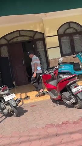 A man wearing a striped polo shirt, dark pants, a baseball cap, and sandals walks towards a building entrance. Two motorcycles, one red and one teal, are parked in front of the building. The ground is paved with reddish-brown tiles. The building appears to be a house or small business with a large wooden door and arched windows. The scene is lit by bright sunlight, casting shadows.