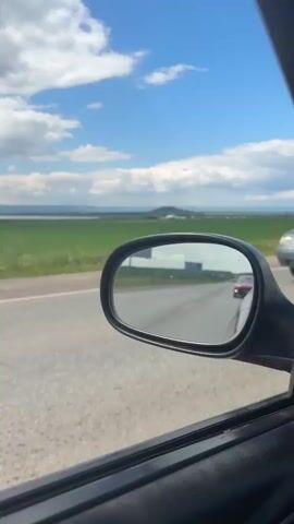 A view from inside a car, looking out the passenger window at the side-view mirror. The mirror reflects a road with several cars driving. The sky is blue with white clouds. Beyond the road, there is a flat, green field. In the distance, there are hills and more clouds.
