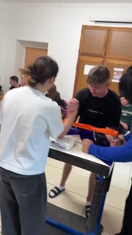 The video shows a group of young people gathered in a room. Several individuals are seated in chairs and watching a pair of young men arm wrestling at a table. A person stands behind the table, observing the arm wrestling match. Flags of various countries are strung across the ceiling. The floor is tiled. Some people in the background are seated in a semi-circle observing the arm wrestling.
