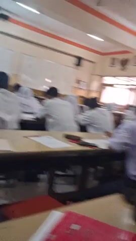 The video appears to be taken from inside a classroom. The perspective is from a student's desk, looking towards the front of the room and then panning to the side. Several students are visible, all wearing white shirts, some with ties. In the foreground, there are various objects on a desk, including what appear to be drinks, papers, and possibly snacks. The walls are painted and have some decorative elements, including what looks like a national emblem. A projector is mounted on the ceiling. The students seem to be engaged in some activity, possibly a lesson or a break. At one point, the camera focuses on a student who smiles directly at the camera. Another student wearing a yellow hijab is visible in the background. The overall atmosphere seems casual and informal.