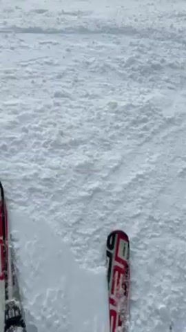 The video shows a snowy scene. The video starts with a close-up of two skis in the snow.  The camera then pans upwards, first showing blue ski poles, and then a landscape with snow-covered mountains and trees under an orange-red sky.