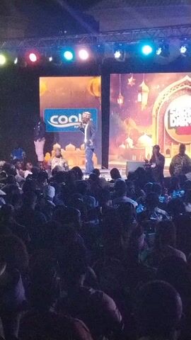 Here's a description of the video:

The scene captures a live stage performance in front of a large audience. A man wearing a suit and a white hat is standing center stage, holding a microphone and addressing the audience. Behind him, two large screens display branding and festive imagery, including a logo for 'Cool Kano'. On stage with him are several other individuals, seated and engaged in what appears to be a panel or discussion.

The audience is densely packed, with many individuals wearing traditional Muslim attire. The lighting is colorful, with spotlights illuminating the stage. Overall, the scene conveys a sense of a well-attended event, likely related to cultural or religious observance.