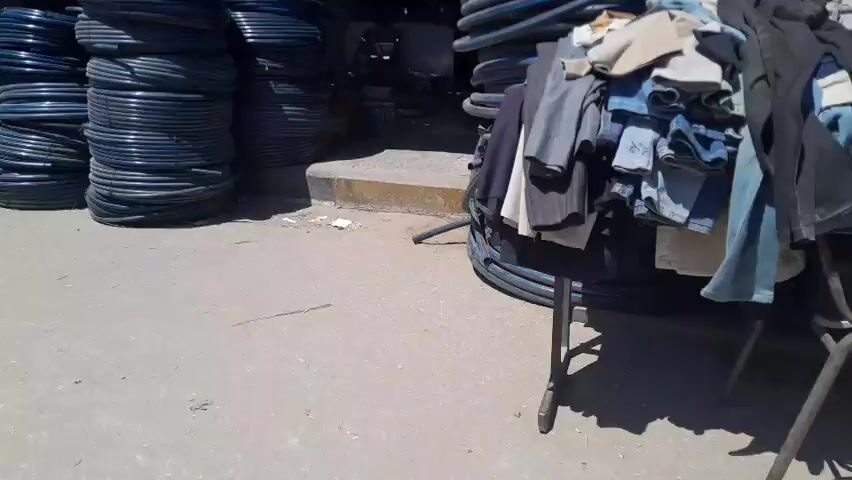 This media file captures a bustling outdoor market scene, likely in Al-Mawasi. The primary subjects are large spools of black piping, piled high and arranged in stacks, dominating the foreground. Amidst these industrial materials, several individuals can be seen. Their clothing suggests a warm, possibly sunny day, with a clear blue sky visible in the background. The setting appears to be a market stall or a vendor's area, with makeshift shelters and some scattered merchandise, including what look like bags of produce and possibly some metal containers.

The activity appears to be the everyday operation of the market. People are moving around, interacting, and likely conducting business related to the sale of the piping and other goods. One person, wearing a blue shirt, is partially visible behind the piping, suggesting they are a vendor or customer. Another individual, with a green shirt, is further back, and a woman with a red top is in the middle ground. The presence of a green bucket and a red bucket hanging from the shelter overhead, along with a sink basin, indicate a functional, working environment.

Notable details include the sheer volume of piping, suggesting it's a primary commodity. The sunlight casts distinct shadows, indicating it's likely midday or early afternoon. The overall impression is one of a busy, working market, with a focus on the sale of goods. There are no obvious signs of distress or unusual events; the atmosphere seems normal for a commercial hub. There is no visible text in the provided frames.