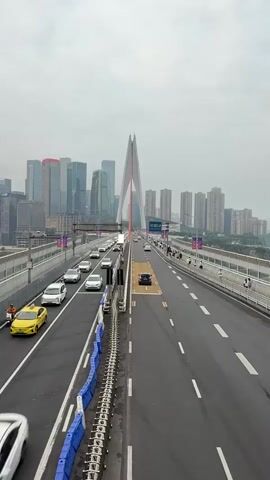 The video shows a vertical panning shot of a multi-lane bridge with moderate traffic. On the left side of the bridge, there are several cars driving in the same direction, including a yellow taxi. A metal railing with blue barriers separates this lane from an adjacent lane with more cars. On the right side of the bridge, there are more cars traveling in the opposite direction. People are visible walking on a pedestrian path on the right side of the bridge. In the background, a city skyline with numerous skyscrapers is visible under a cloudy sky. The bridge itself features a prominent white cable-stayed tower in the center. The overall environment appears to be urban and busy.