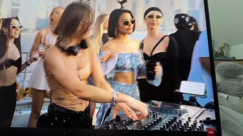 The media file shows a group of people on what appears to be a boat or a large deck, with a cityscape in the background. The scene suggests a party or gathering. In the foreground, a person is operating a DJ mixer, suggesting music is playing. Several women are visible, dressed in party attire, some holding drinks. One woman is wearing a bikini top and a white skirt, another a blue and white gingham off-the-shoulder top and skirt. Others are in dresses and fashionable outfits.

The time of day appears to be daytime, with bright lighting. The weather seems to be clear. The backdrop suggests an urban environment with tall buildings, possibly a waterfront. The mood seems festive and energetic, with people engaged and enjoying themselves.

There is no visible text in the media file. The specific location of Falenty Nowe, Poland, is not visually discernible from the provided image.