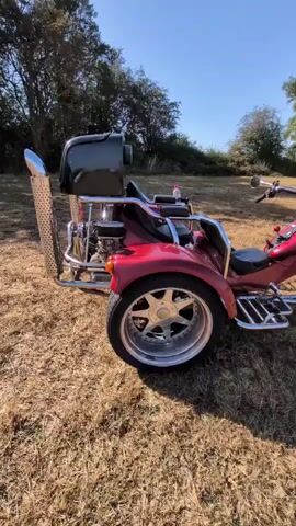The video displays a close-up, panning view of a red three-wheeled motorcycle, or trike, parked on dry grass in an outdoor, rural setting. The trike features chrome accents, including large exhaust pipes, decorative trim, and wheels. The camera focuses on the rear of the vehicle, then pans to show the front, revealing two seats and handlebars. The background consists of trees and a clear blue sky, indicating it is daytime. No people are visible in the video, and the trike appears stationary.