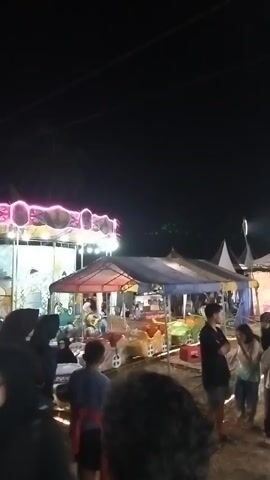 The video shows a night-time outdoor event with several tents and amusement rides. A carousel with bright pink lights is visible on the left side of the frame. In the center, a smaller ride with colorful seats is covered by a blue and red tent, with people standing around it. To the right, more tents are set up, and a crowd of people, many wearing hijabs, are milling about. The ground appears to be dirt. The overall atmosphere suggests a festive gathering or fair.