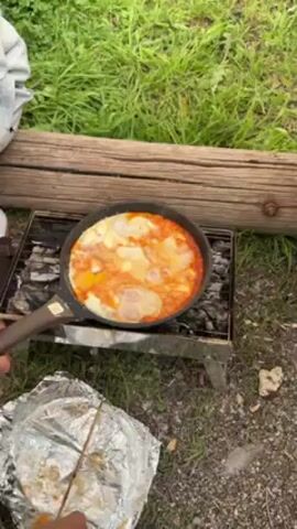 The video shows a frying pan being used to cook eggs in tomato sauce over charcoal embers outdoors. The pan sits on a small, foldable grill. The background consists of green grass and a log.  A person's hand is visible near a piece of foil-covered food on a stick. The scene is static with the camera angle remaining constant.