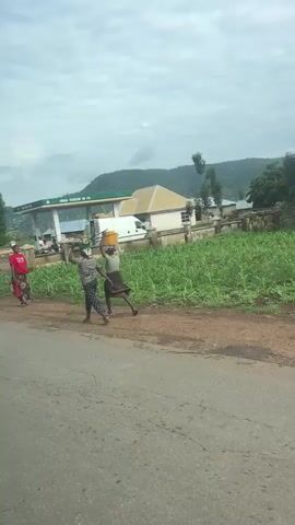 The video shows a view from a moving vehicle. The first scene captures a gas station with a green awning and pumps. A white van is parked to the right, and several people are visible near the station. A woman in a red shirt and patterned skirt walks away from the camera on a dirt path. To the left of the gas station, there are buildings and a fence. A field of green crops extends to the right. In the distance, green hills rise under a cloudy sky. The second scene shows a person standing in the middle of the gas station lot, holding up a white object. The third scene shows a large tree and a few people sitting on a low wall. The final scenes continue to show the green fields and hills, with a woman carrying a yellow container on her head walking along the side of the road.