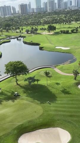 This is an aerial drone shot of a golf course with a pond and several sand traps. The golf course is surrounded by palm trees and other vegetation. In the background, there are tall buildings, suggesting an urban environment. There is a golf cart visible on the course, and a person is seen on the green. The sun is casting long shadows, indicating it is either early morning or late afternoon.
