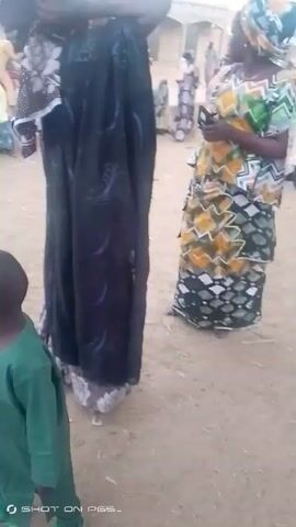 The video shows an outdoor scene in what appears to be a village or small town. Several people are seen wearing traditional clothing. Two women are standing in the foreground, one dressed in a black garment and the other in a colorful patterned dress. As the camera moves, it reveals more people in the background, including men and children, and some are sitting on the ground. The ground is sandy. There are trees, buildings, and some vehicles visible in the background, suggesting it could be a public gathering or a market area.