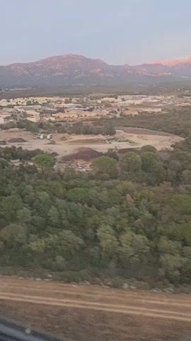 The video, recorded from an aircraft window, shows an aerial view of a rural landscape at dusk. In the foreground, there is a patch of dry earth and some vegetation. Beyond that, a dense green forest occupies the mid-ground. Further back, a town or industrial area is visible, characterized by numerous buildings, roads, and construction sites with machinery. In the distance, a range of mountains is silhouetted against the pale evening sky, with the peaks catching the last rays of sunlight. The camera perspective is static, and no individuals or direct interactions are discernible. The overall scene depicts a transition from natural wilderness to human development, set against a mountainous backdrop.