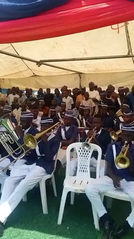 Under a tent with red and blue fabric draped above, a brass band comprised primarily of young people plays instruments like trumpets, trombones, and tubas. They are dressed in matching navy blue shirts, white pants, and striped hats. They sit on white plastic chairs, arranged in rows. An audience of adults and children watch. The setting seems to be outdoors on artificial turf.