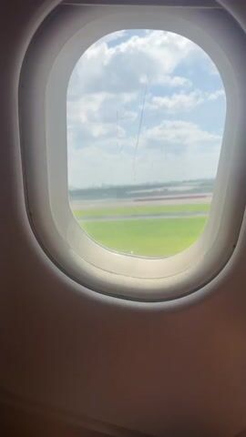 The video shows a view from an airplane window as the plane is on the runway preparing for takeoff or landing. The window is oval-shaped with a white frame. Outside the window, a green grassy field and a runway are visible under a partly cloudy sky. A city skyline can be seen in the distance. The window pane has a few thin cracks. Towards the end of the video, a hand holding a smartphone enters the frame from the bottom right corner. The phone's screen is dark.