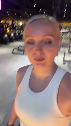 A woman with short blonde hair and light makeup, wearing a white tank top and black workout gloves, is in a gym. She is looking at the camera and smiling. In the background, there are rows of exercise machines, including stationary bikes and weight machines. The gym appears to be dimly lit with purple accent lighting. Other people can be seen in the background, some of whom are using the equipment. The camera is focused on the woman's face.