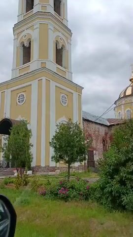 The video pans from left to right, showing a bell tower and a church. The bell tower is tall and slender, painted yellow and white, with arched openings. To the right of the bell tower is a church with a golden dome. The church is made of red brick and has windows. In front of the buildings, there is a garden with green grass, small trees, bushes, and pink flowers. The sky is cloudy. A red brick archway is visible in the foreground on the left.