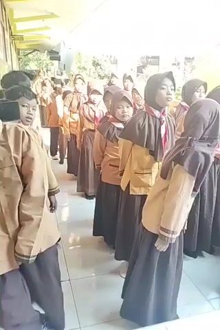 A group of school children in uniform are standing in a line outside a building. The uniforms consist of a brown shirt, a tan vest, and a dark brown skirt or pants. Some of the girls are wearing headscarves. They appear to be in an outdoor corridor or walkway. The children are looking forward, and some appear to be listening to someone. The lighting suggests it is daytime.