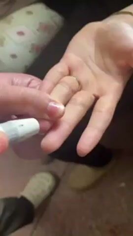 The video shows someone pricking the finger of another person to draw blood. The person doing the pricking is holding a white, pen-like device and pressing it against the pinky finger of the other person's hand. A small drop of blood appears on the fingertip after the prick. The person receiving the prick is wearing a ring on their ring finger. The background includes a cushion with a floral pattern, black pants, and beige shoes with polka dots.