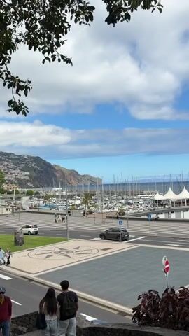 The video shows a marina with many sailboats docked, a road with crosswalks, and a few pedestrians walking across. There is a building near the marina with rows of stadium seats in front. A car drives on the road. The sky is blue with white clouds. The camera pans slightly to the right.