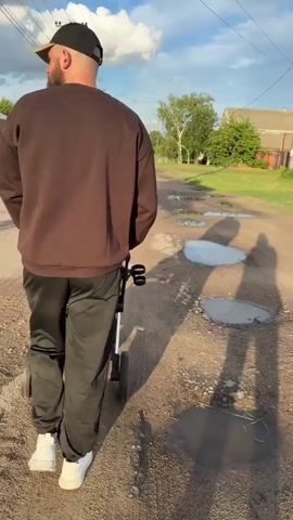 A man with a beard and a black baseball cap is walking away from the camera down a dirt road. He is wearing a brown sweatshirt and black pants. He is pushing a stroller. The road is lined with trees and houses. There are puddles of water on the road, suggesting it has recently rained. The sun is setting, casting long shadows. The sky is blue with some white clouds. The overall impression is one of a peaceful evening stroll in a rural setting. There is no visible text in the media file.